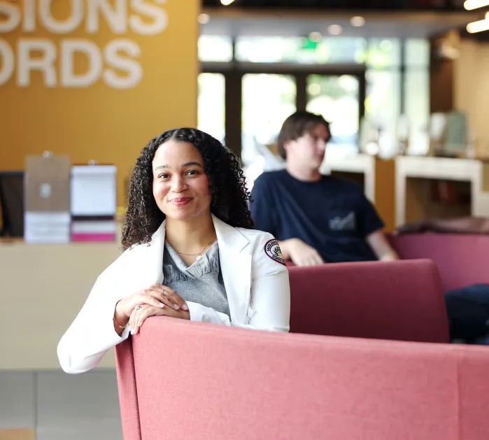 Female student sitting on couch in lobby with other students talking in the background. 