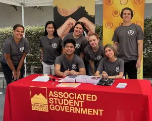 Seven students representing the Associated Student Government pose and smile for a photo while standing behind a table with a red table cloth that reads Associated Student Government