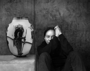 Black and white image. Artist Virgil Ortiz sits, in a black shirt and jeans with his hands together, next one of his pieces, which is an oval-shaped piece of pottery featuring a Native American warrior. 