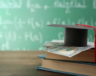 A stack of textbooks, with a few $100 dollar bills on top of it and a mortar board resting on top of the money. 