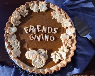A pumpkin pie decorated with pastry cutouts spelling “Friendsgiving” in the center and surrounded by leaf- and pumpkin-shaped crust pieces along the edge. The pie sits on a blue cloth with a metal pie server beside it.