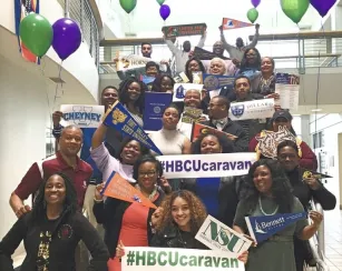 A large, joyful group of people of various ages stands on indoor stairs holding signs and banners representing Historically Black Colleges and Universities (HBCUs). The group displays school pennants for institutions such as Florida A&M, Dillard, Fort Valley State, Cheyney, and Bennett College, along with large signs reading “#HBCUcaravan.” Colorful balloons in green and purple are tied above the group, and the photo is taken in a bright, modern building.