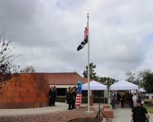A Veterans Day ceremony at Saddleback College shows uniformed officers saluting as the American and POW/MIA flags are raised on a flagpole. People stand under white canopies watching the event, and red, white, and blue balloon decorations are nearby. The ceremony takes place in front of a curved copper memorial wall under a cloudy sky.