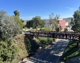 A pedestrian bridge with large metal letters spelling “Saddleback College” spans a campus road. Trees and greenery surround the bridge, and in the background, a red and white campus building sits against rolling hills under a clear blue sky.