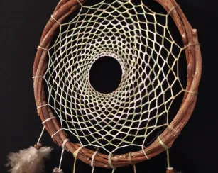A handcrafted dreamcatcher made with a circular wooden hoop wrapped in twine, featuring an intricate woven web pattern with a hole in the center. Several soft brown and white feathers hang from the bottom, each tied with a small wooden bead. The background is black, highlighting the details.