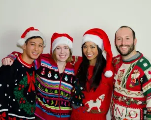 Four adults stand close together, smiling at the camera and wearing festive holiday sweaters and Santa hats. Their sweaters include patterns such as bows, trees, snowmen, animals, and the word “NO.” They pose against a plain background.