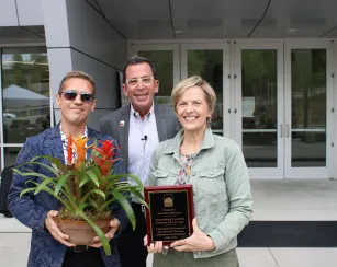 Three adults stand smiling outside a building entrance. The person on the left holds a potted plant with long leaves and tall flower spikes. The person in the center stands behind them, wearing glasses and a microphone. The person on the right holds a plaque that reads: “Presented To Marlene Nielsen. Outstanding Emeritus Professor of the Year. In Recognition Of Exceptional And Influential Teaching, Commitment, And Dedication. 2004–2023.”
