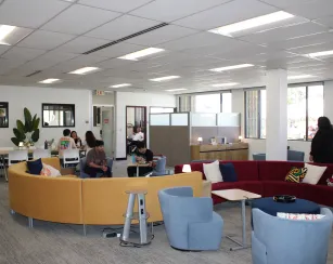 A spacious student lounge area with groups of people sitting and talking at small tables and on curved sofas. Several chairs, tables, and workstations are arranged throughout the room. A few students are studying or using laptops, while another person stands near the windows. Large windows line the back wall, and enclosed offices or meeting rooms are visible along the left and far walls. There is no visible text in the image.
