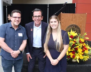 Three adults stand smiling in front of a small stage at a Saddleback College event. A podium with the Saddleback College seal and a large yellow-and-red floral arrangement are behind them, with a microphone set up on stage.