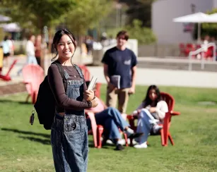 Female Saddleback College student standing with backpack and book with on the quad with students sitting in group in the background.