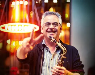 A smiling man with short gray hair and glasses stands outdoors at night, holding a saxophone in one hand and flashing a peace sign with the other. Warm orange lights glow behind him, including a circular neon sign that reads “AMORE,” creating a vibrant, blurred background.