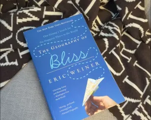A blue paperback book titled “The Geography of Bliss” by Eric Weiner rests on a gray surface beside a black-and-cream patterned blanket. The cover reads: “The New York Times Bestseller,” “One Grump’s Search for the Happiest Places in the World,” “The Geography of Bliss,” “Eric Weiner,” “Charming, funny, and illuminating. – Washington Post Book World,” and “Moving, profound… a winning book. – National Geographic.” The cover features a hand holding a paper airplane made from a map.