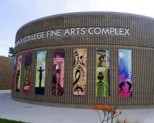 Curved brick building with large metal letters reading “Saddleback College Fine Arts Complex.” Seven vertical banners on the wall read, from left to right: “Dance,” “Art,” “Speech,” “Entertainment & Theatre Technology,” “Music,” “Art History,” and “Photography.” A walkway and small plants are in front of the building.