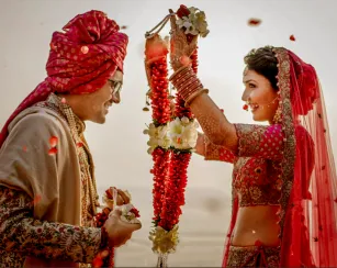 A bride and groom face each other during a wedding ceremony. The bride, wearing a red embroidered outfit with jewelry and a veil, lifts a flower garland to place around the groom’s neck. The groom, wearing a patterned outfit and a red turban, smiles and holds a matching garland. Flower petals fall around them.