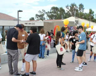A lively outdoor student event on a college campus plaza, with diverse groups of young adults talking, holding papers, and carrying matching Saddleback College tote bags in front of a "Fine Arts (FA)" building.