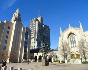 A sunny view across Boston University's Marsh Plaza, featuring the Gothic-style Marsh Chapel, a central bronze sculpture, and campus high-rises under a clear blue sky.