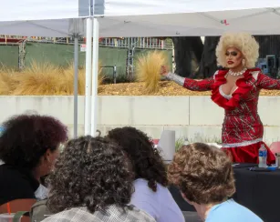 A drag queen with voluminous blonde hair and a red, silver, and black sequined dress gestures with outstretched arms while hosting an outdoor bingo event beneath a white canopy. Audience members watch from tables in the foreground.