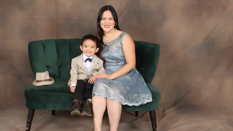 A studio portrait of a smiling woman in a light-blue lace dress and her young son in a tan blazer and bow tie, sitting together on a green velvet couch against a brown backdrop.
