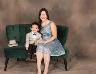 A studio portrait of a smiling woman in a light-blue lace dress and her young son in a tan blazer and bow tie, sitting together on a green velvet couch against a brown backdrop.