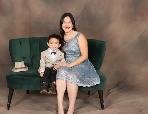 A studio portrait of a smiling woman in a light-blue lace dress and her young son in a tan blazer and bow tie, sitting together on a green velvet couch against a brown backdrop.