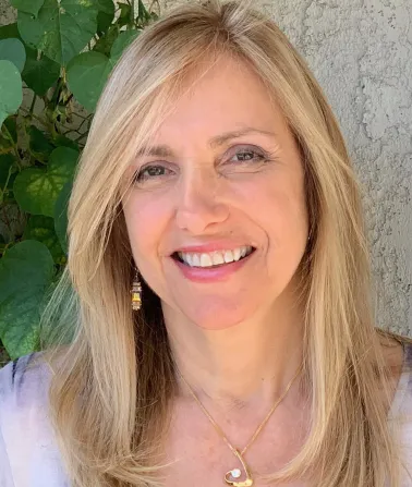 A woman with long blonde hair smiling in front of a neutral wall and green leafy plants. She is wearing a light-colored top, a gold necklace with a pendant, and a single dangling earring.