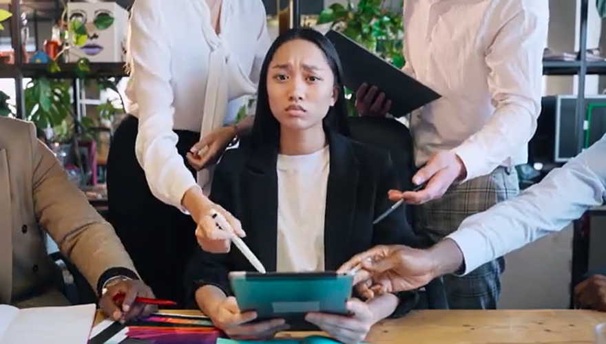 Female student sitting at a desk looking haggard with many people around her pointing at things causing chaos.